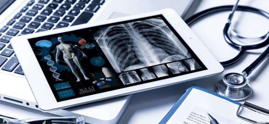 Ein Tablet liegt auf einer Laptoptastatur und zeigt Röntgenaufnahmen und medizinische Darstellungen von Organen, Knochen und DNS. Daneben liegt ein Stethoskop.