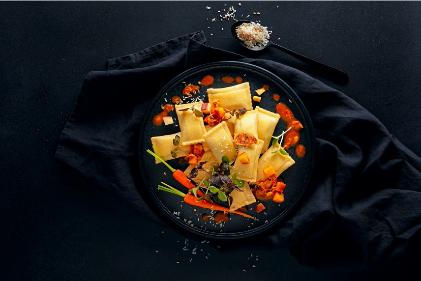 auf einem schwarzen Hinterrund liegen ein Löffel und eine Kochschürze, auf der ein Gericht mit Maultaschen und Karotten zu sehen ist.
