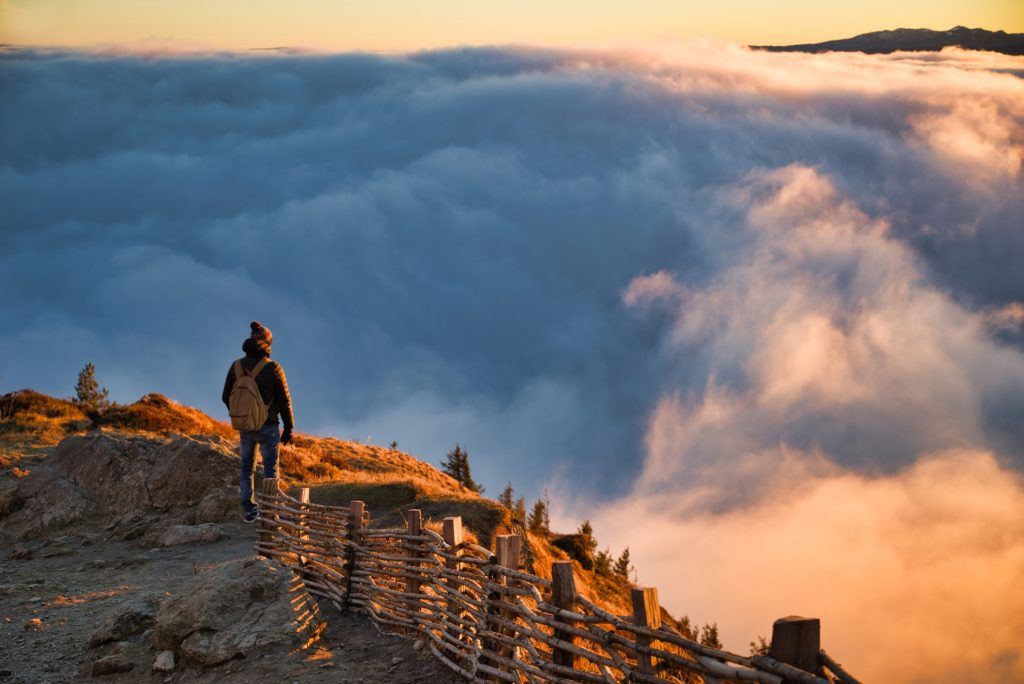 Mann steht auf einem Berg und schaut in der Abendsonne auf die Wolken, die sich unter ihm gesammelt haben.