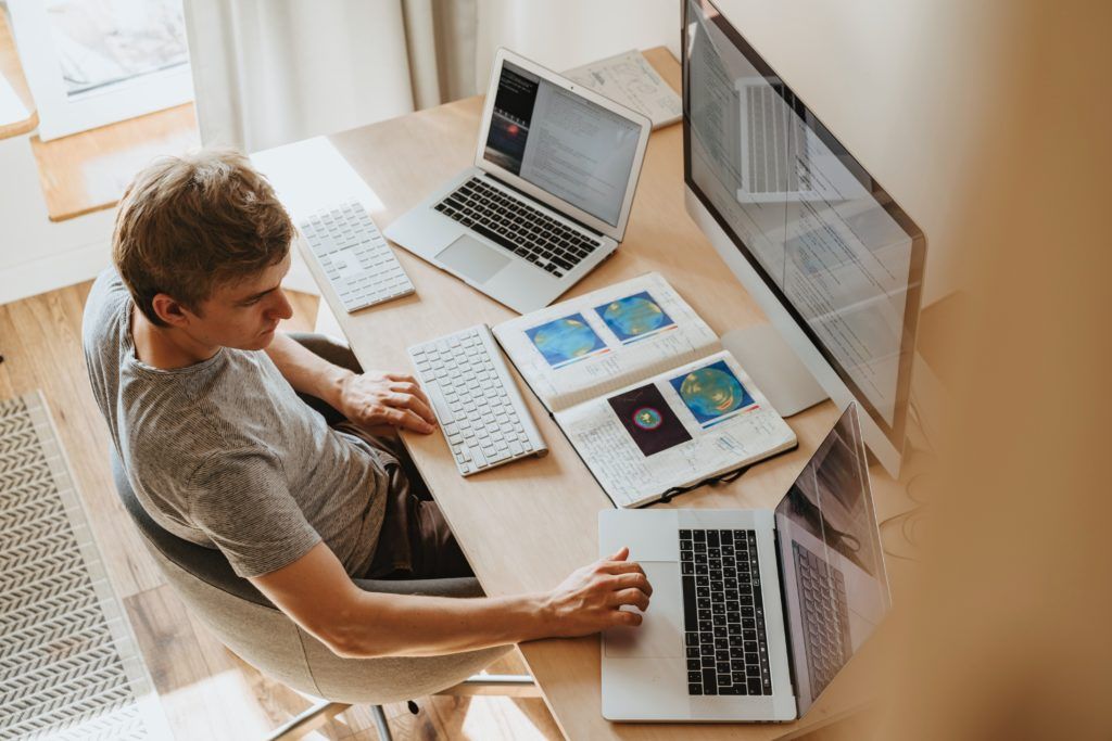 Ein Mann sitzt am Schreibtisch vor zwei Laptops, Bildschirm und Notizbuch