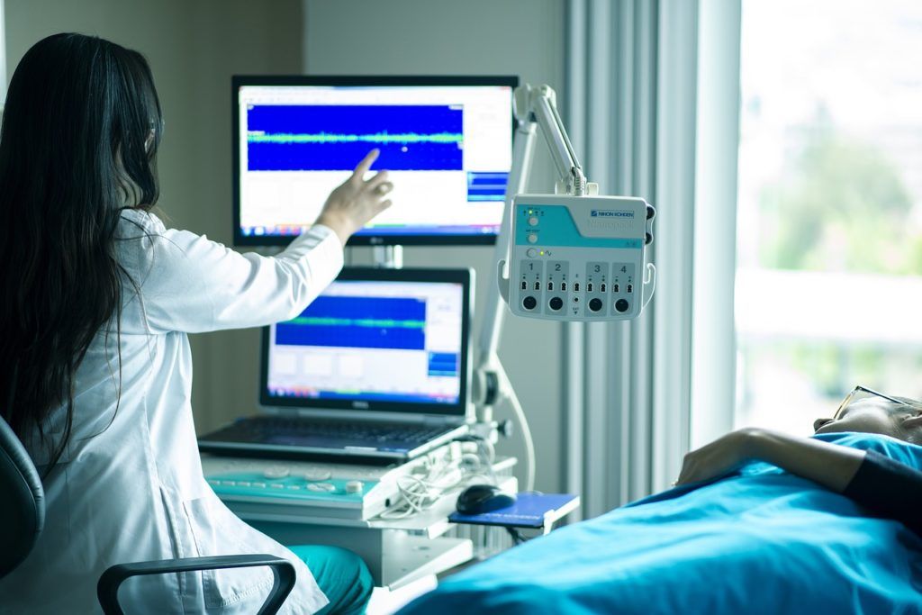 Frau auf Krankenliege. Ärztin zeigt auf einen der zwei Bildschirme.